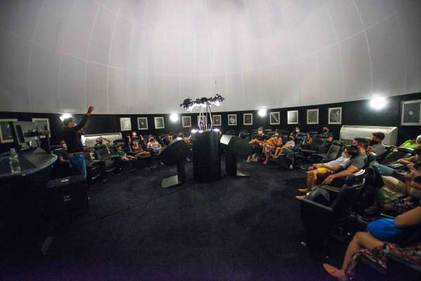 Fotografia em ambiente interno. Imagem ampla de dentro do planetário com pessoas sentadas em cadeiras em volta de aparelho ao centro da sala. À esquerda, há um homem em pé com o braço direito levantado. Na parte superior da fotografia, percebe-se a cúpula do planetário.